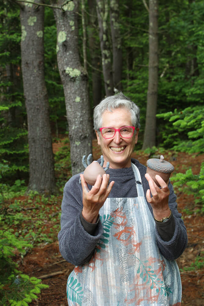 A smiling artist holds two ceramic sculptures in the woods